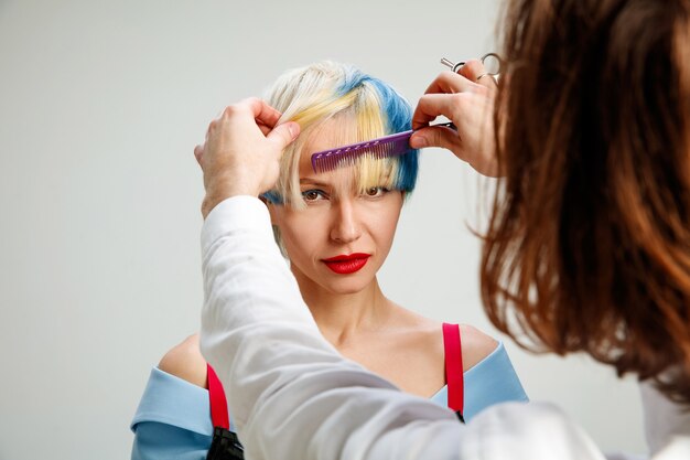 Foto mostrando uma mulher adulta no salão de cabeleireiro. Foto de estúdio de graciosa jovem com corte de cabelo curto elegante e cabelo colorido em fundo cinza e mãos de cabeleireiro.