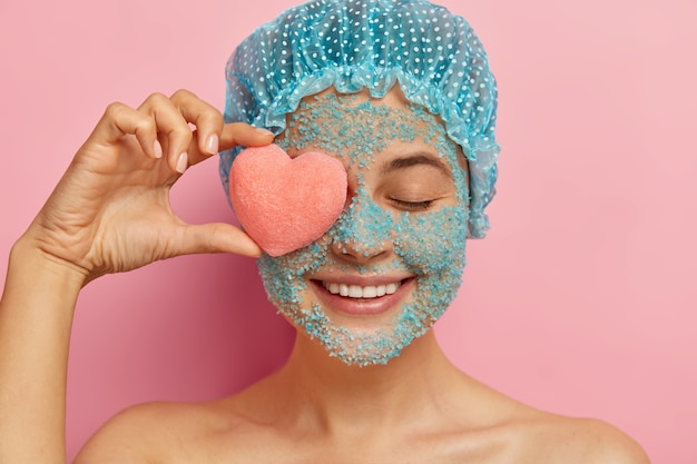 Foto grátis foto na cabeça de uma jovem alegre com esfoliante de sal marinho cristalino, segurando uma esponja em forma de coração rosa no olho, sorrindo positivamente, usando uma capa de chuva, modelos contra uma parede rosa, descascando o rosto dos poros