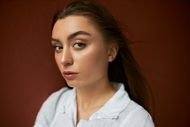 Foto na cabeça de uma jovem atraente e elegante com uma maquiagem elegante, pele perfeita e cabelos castanhos compridos, olhando para a câmera com expressão facial séria, vestindo uma camisa formal branca, com aparência confiante