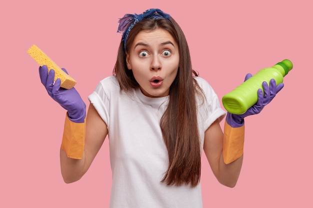 Foto na cabeça de uma jovem caucasiana surpresa, olhando maravilhada, usando roupas casuais, segurando um pano e um frasco de detergente