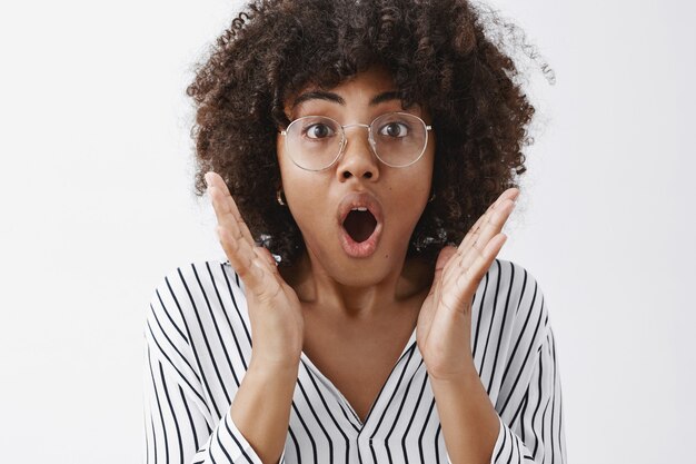 Foto na cabeça de uma mulher afro-americana atraente e impressionada com um penteado afro em óculos transparentes, arfando, abrindo a boca de espanto e choque, de mãos dadas perto do rosto em pé tremeu sobre a parede cinza