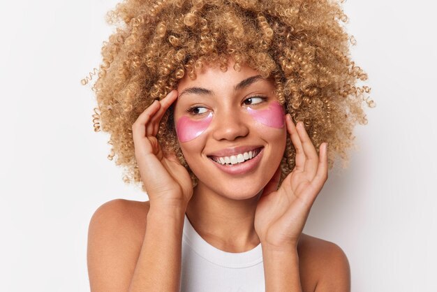 Foto na cabeça de uma mulher bonita e positiva tocando o rosto, parece feliz, longe, tem uma expressão gentil, usa almofadas de colágeno rosa sob os olhos, sente-se satisfeita, cuida da pele isolada no fundo branco