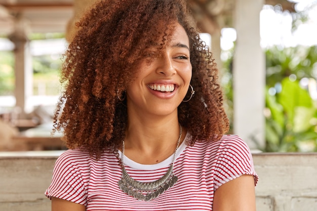 Foto na cabeça de uma mulher negra engraçada com cabelo encaracolado, ri de uma boa piada, tem um sorriso cheio de dentes, mostra dentes brancos perfeitos, usa uma camiseta listrada casual, posa na lanchonete do terraço