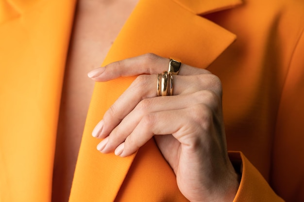 Foto próxima de uma mão fina e graciosa de uma garota com uma manicure na moda primavera nua e um anel de ouro nos dedos em uma jaqueta laranja