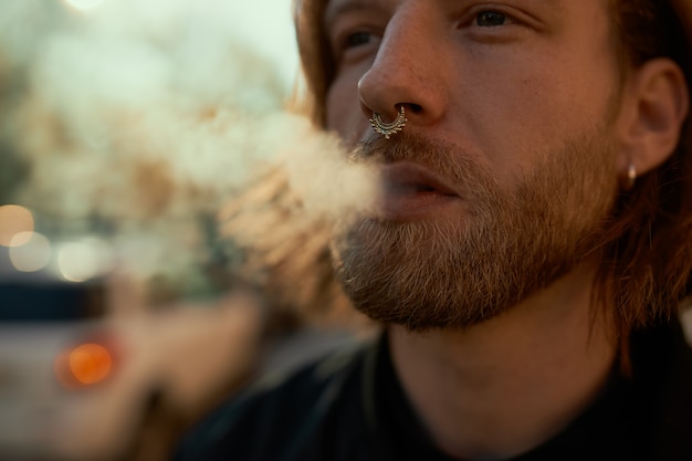 Foto grátis foto recortada de cigarro eletrônico de vaping masculino barbudo elegante jovem bonito ao ar livre. perto de um cara atraente com argola no nariz soltando fumaça enquanto caminhava na rua em um dia ensolarado de verão