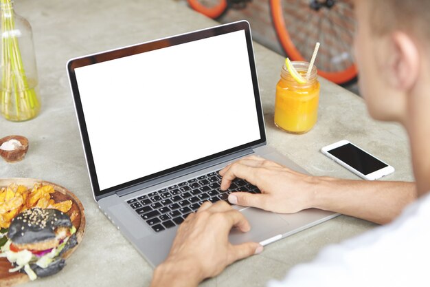 Foto recortada de jovem freelancer usando laptop para trabalho distante, sentado na mesa de café com telefone celular de tela em branco