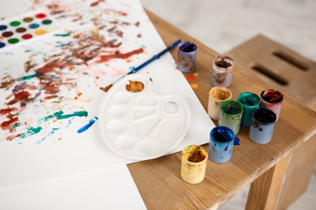 Foto recortada de mesa de madeira com tintas, pincéis, paleta e fotos feitas por crianças
