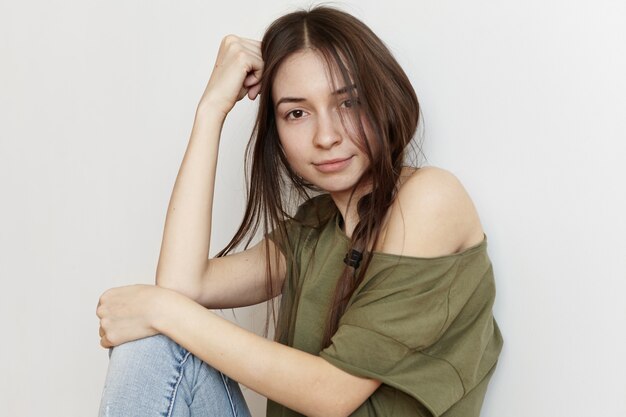 Foto recortada de moda feminina jovem bonita com penteado desarrumado, vestindo roupas elegantes, descansando a cabeça no cotovelo e sorrindo, um ombro aparecendo em sua camiseta de tamanho grande do exército verde