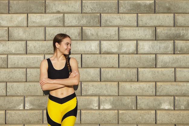 Foto recortada de uma jovem linda e positiva vestindo roupas esportivas pretas e amarelas da moda, descansando ao ar livre, posando contra uma parede de tijolos em branco com espaço de cópia para seu conteúdo