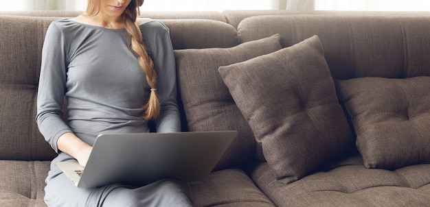 Foto recortada de uma jovem loira com uma trança trabalhando em um laptop sentado no confortável sofá escuro em casa, luz quente retroiluminada. Conceito de freelance ou estilo de vida.