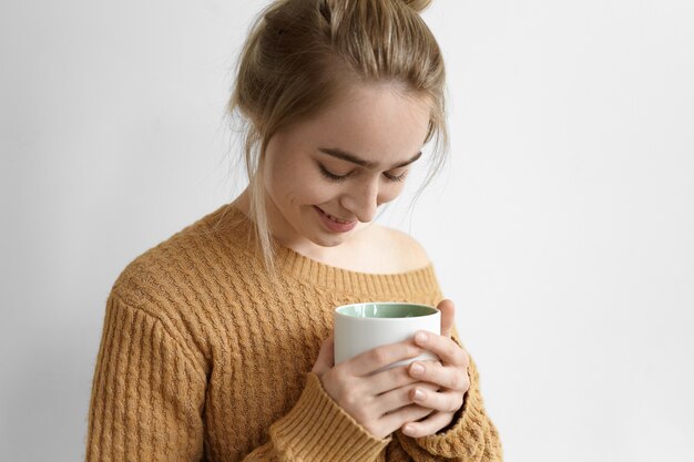 Foto recortada de uma linda jovem europeia vestida casualmente com coque de cabelo posando com uma camisola enorme, segurando a caneca com as duas mãos, olhando para baixo e sorrindo timidamente. Descanso e relaxamento