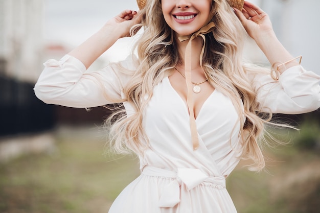 Foto recortada de uma linda mulher loira com cabelo longo ondulado, usando chapéu com laços e vestido branco. Fundo desfocado.