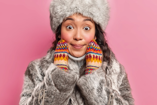 Foto recortada de uma mulher de inverno atônita em agasalhos quentes olhando para a frente de forma surpreendente, mantendo as mãos no rosto em poses contra a parede rosa