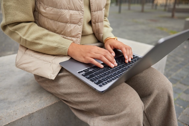 Foto recortada de uma publicação de texto irreconhecível do tipo blogueira feminina em um laptop portátil funciona remotamente ao ar livre, vestida com roupas casuais, teclados em um dispositivo moderno se prepara para os exames