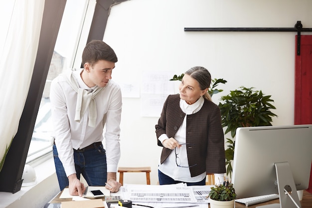 Foto sincera de dois arquitetos europeus discutindo no escritório, em pé na mesa com computador, desenhos e ferramentas, sorrindo um para o outro, satisfeitos com o trabalho comum. Pessoas e cooperação