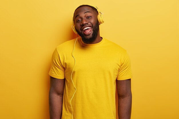 Foto sincera de um homem afro-americano feliz curtindo um podcast de música e usando fones de ouvido estéreo modernos