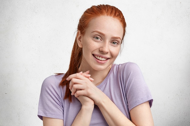 Foto sincera de uma aluna satisfeita e satisfeita, alegra-se com seu sucesso durante os estudos, sorri gentilmente para a câmera, tem o cabelo ruivo preso em um rabo de cavalo, vestida com uma camiseta roxa casual