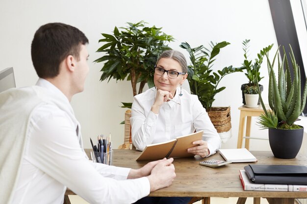 Foto sincera de uma CEO feminina de meia-idade positiva e atraente usando óculos, fazendo anotações no caderno, ouvindo um jovem candidato a emprego durante uma entrevista em seu escritório aconchegante