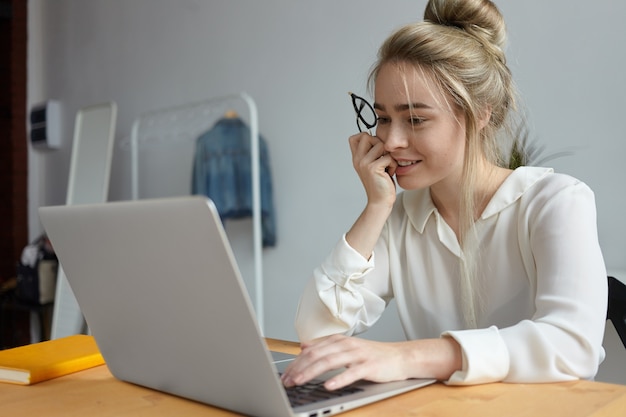 Foto sincera de uma jovem freelancer positiva com um penteado bagunçado usando um dispositivo eletrônico portátil genérico na mesa de madeira, trabalhando remotamente do escritório em casa, tendo absorvido o olhar interessado