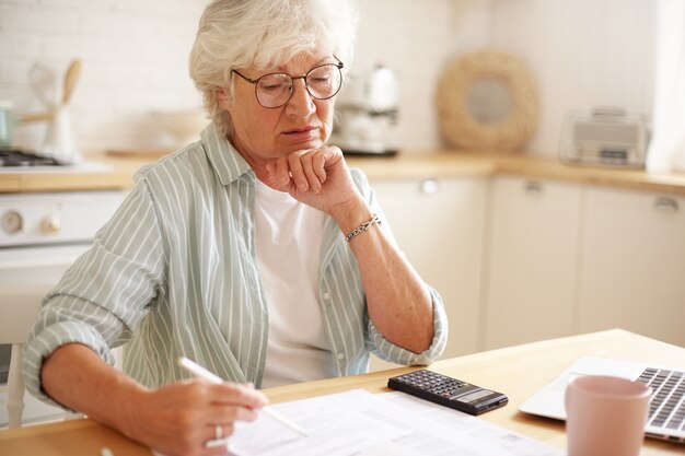 Foto sincera de uma mulher caucasiana aposentada séria usando óculos, calculando despesas, tentando economizar dinheiro para uma compra cara, pagando contas domésticas online usando um dispositivo eletrônico na mesa da cozinha