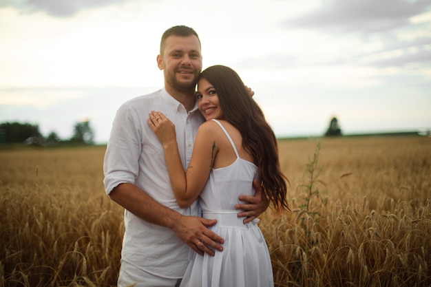 Foto stock de comprimento total de um casal romântico em roupas brancas, abraçando-se no campo de trigo ao pôr do sol.