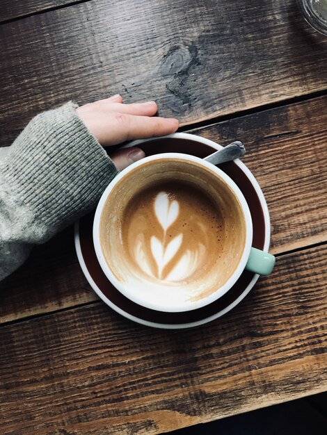 Foto vertical aérea da mão de uma pessoa perto do café latte art em uma superfície de madeira