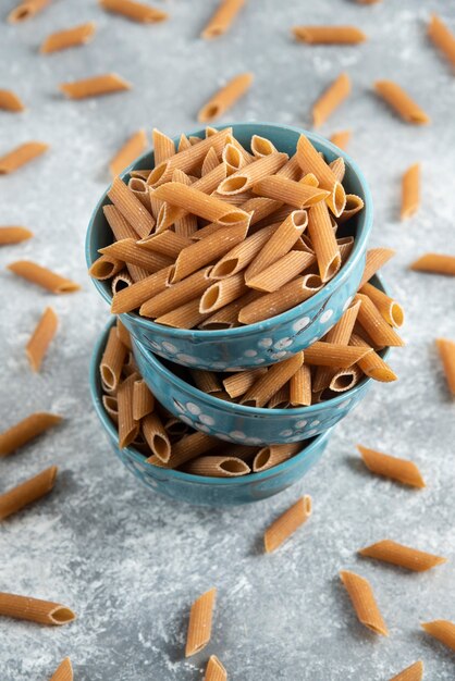 Foto vertical da pilha de tigelas cheias com macarrão penne dietic marrom sobre uma superfície cinza.