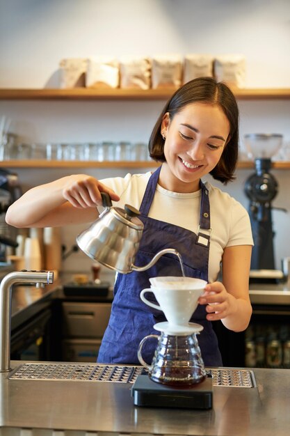 Foto vertical de barista asiático sorridente em avental azul derramando água com uma pequena chaleira fazendo café atrás do balcão em seu café