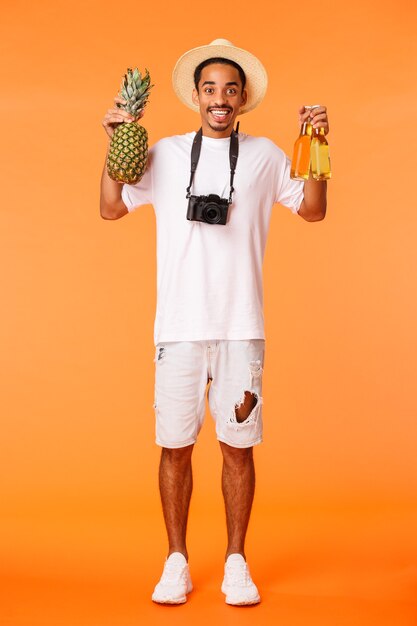 Foto vertical de corpo inteiro, cara afro-americano feliz, sugerindo pegar uma bebida e curtir as férias, segurando abacaxi e duas garrafas de cerveja, sorrindo entusiasmado, relaxando no lazer, fundo laranja