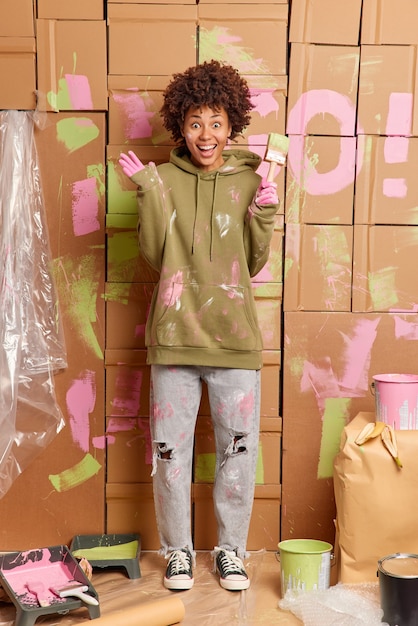 Foto vertical de feliz mulher de pele escura cores paredes do apartamento levanta mãos segura pincel usa moletom sujo e jeans cercado com ferramentas de pintura renova casa após a mudança