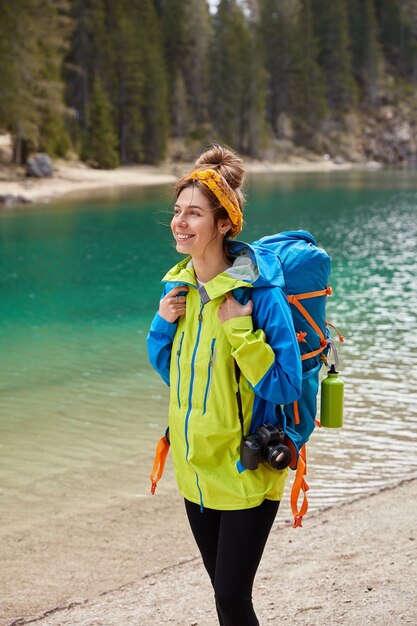 Foto vertical de garota turista caminhando pelo lago turquesa, floresta de coníferas, sorrindo alegremente segurando a câmera e uma mochila grande