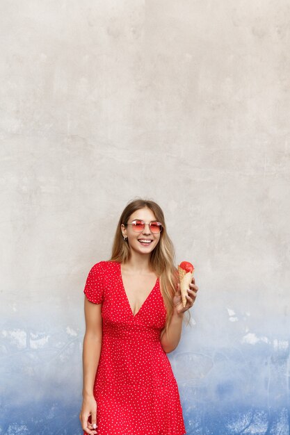 Foto vertical de garota verão tomando sorvete perto da parede da rua e sorrindo para a câmera