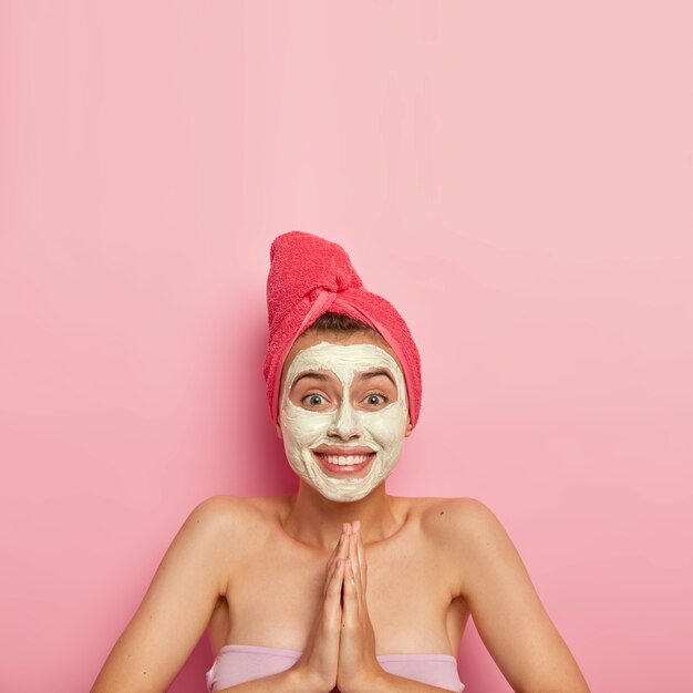 Foto vertical de jovem feliz aplica máscara de limpeza no rosto, mantém as palmas das mãos pressionadas, pede ajuda, recebe tratamentos de beleza, usa toalha enrolada na cabeça, pele saudável, se preocupa com a higiene
