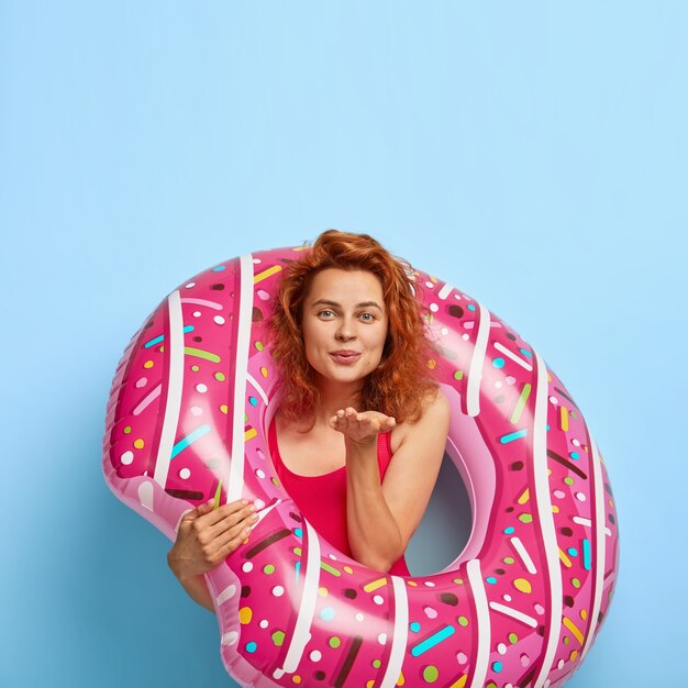 Foto vertical de modelo feminino atraente mandando beijo no ar, vestida de maiô, fica com donut de borracha, expressa amor, tem férias de verão