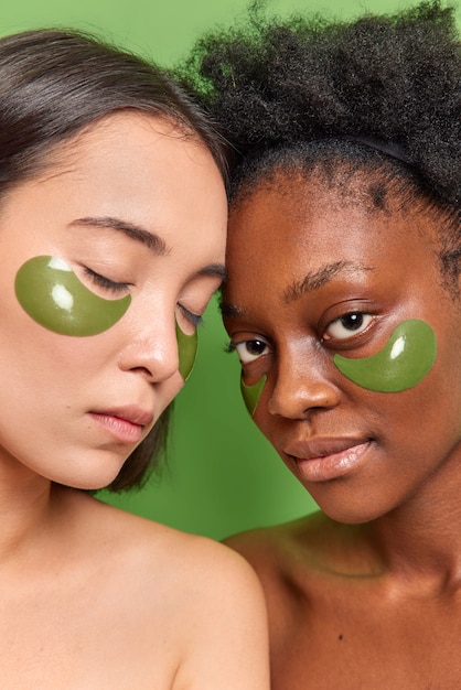 Foto grátis foto vertical de mulheres com pele diferente, ombros nus contra uma parede verde vívida aplique adesivos de hidrogel sob os olhos para reduzir as linhas finas, passe por tratamentos de beleza. cuidados com a pele