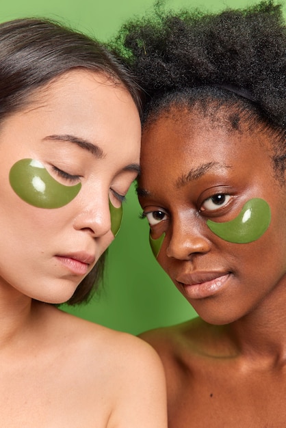 Foto vertical de mulheres com pele diferente, ombros nus contra uma parede verde vívida aplique adesivos de hidrogel sob os olhos para reduzir as linhas finas, passe por tratamentos de beleza. Cuidados com a pele