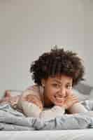 Foto grátis foto vertical de senhora de aparência agradável satisfeita com cabelos cacheados de sorriso cheio de dentes, deitada de bruços em uma cama confortável, tem fim de semana, desfruta de conforto e ambiente doméstico aconchegante. conceito de pessoas e descanso