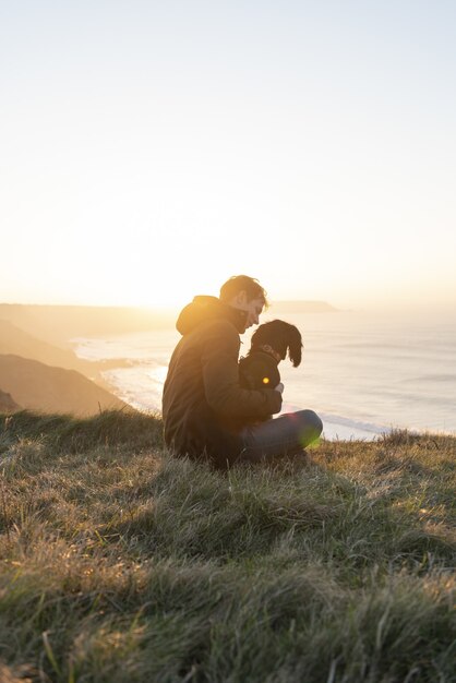 Foto vertical de um homem branco apreciando o pôr do sol no oceano com seu cachorro