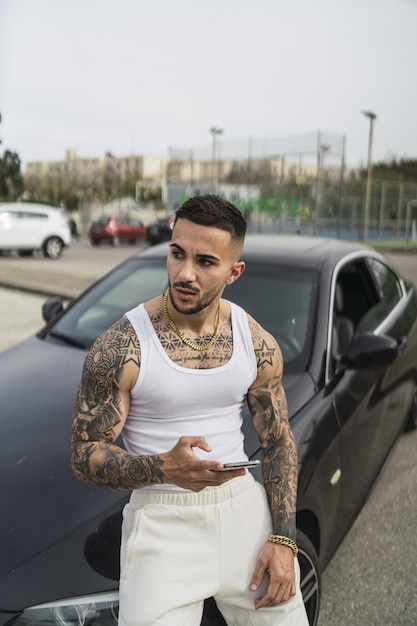 Foto grátis foto vertical de um homem sexy tatuado usando seu telefone encostado em seu carro