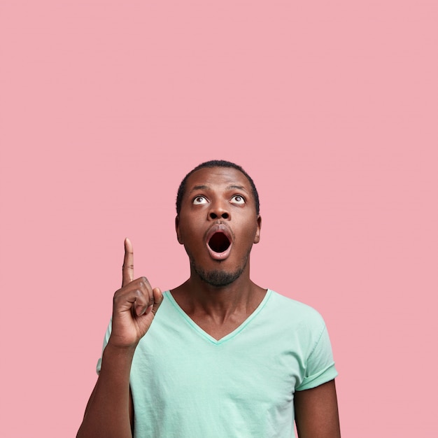Foto vertical de um jovem afro-americano espantado com a boca bem aberta e uma expressão inesperada, vestido com uma camiseta casual, isolado sobre rosa