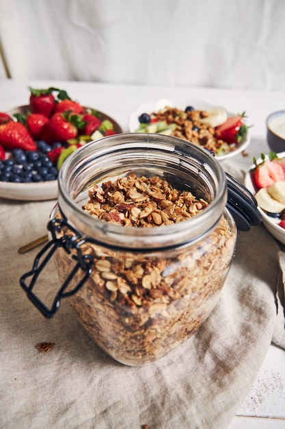 Foto vertical de um pote de granola ao lado de tigelas de frutas, frutas vermelhas e iogurte