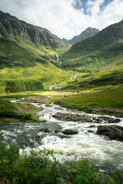 Foto vertical de um rio cercado por montanhas e prados na Escócia