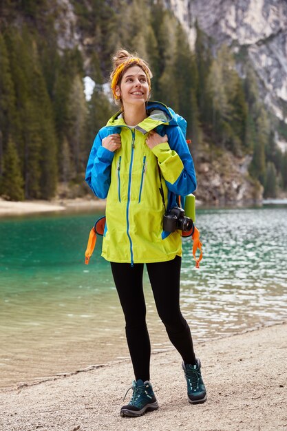 Foto vertical de um viajante positivo caminhando pela costa perto do lago da montanha, carregando uma câmera