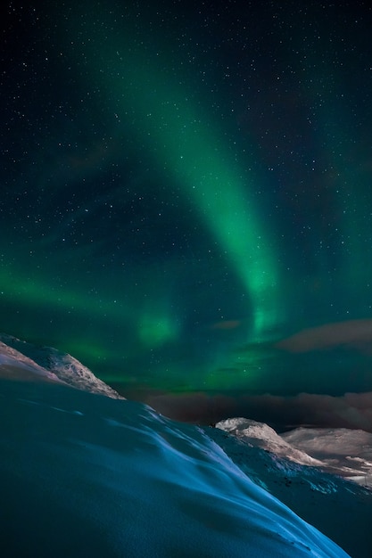 Foto vertical de uma aurora no céu acima das colinas e montanhas cobertas de neve na Noruega