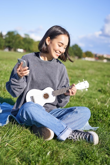 Foto vertical de uma garota coreana sorridente sentada no parque com ukulele e smartphone usando aplicativo móvel para