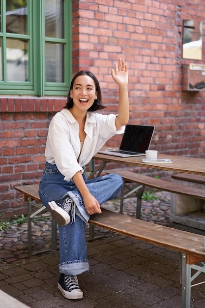 Foto grátis foto vertical de uma garota feliz atraindo atenção acenando com a mão para um amigo no café sentado no banco com