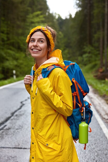 Foto vertical de uma jovem europeia sorridente e feliz usando uma bandana amarela, capa de chuva e carregando mochila