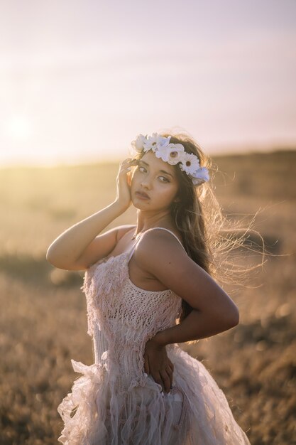 Foto vertical de uma jovem mulher branca com vestido branco e grinalda de flor branca posando em um campo