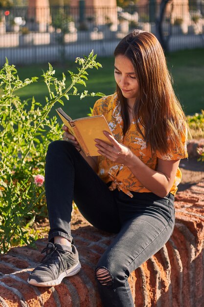 Foto vertical de uma menina em uma camisa amarela lendo um livro sentada ao lado de plantas