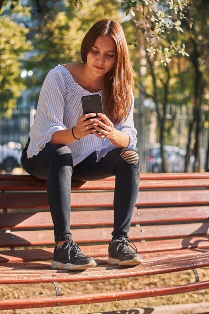 Foto vertical de uma menina sentada no banco olhando para o telefone